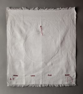 A white cloth hanging up, with an embroidered white circle in the middle, and the words embroidered in red, eat, love, play, bleed in a row at the bottom.